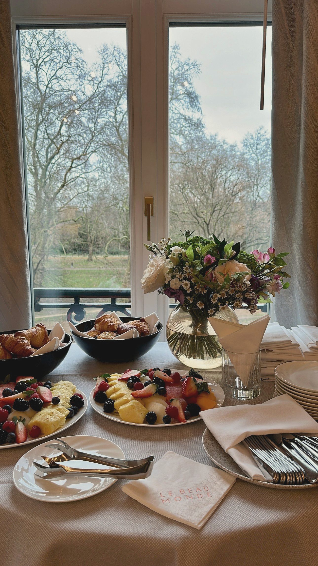 A breakfast spread at Le Beau Monde, laid by a tall window: croissants and pastries, a fruit plate, cutlery wrapped in linen, bare winter trees and a park beyond the glass