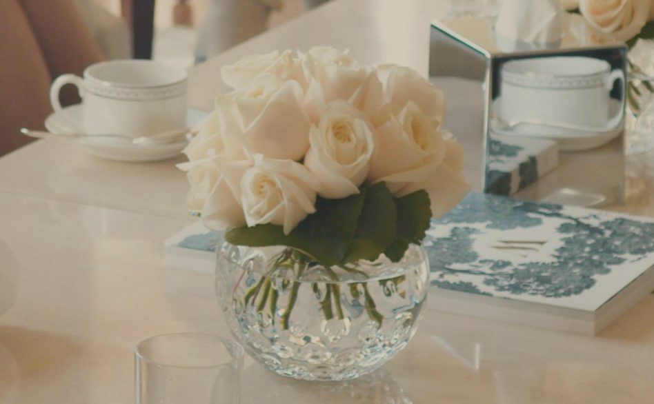 White roses in a crystal bowl beside gold-rimmed porcelain and a toile-bound book, on marble in daylight