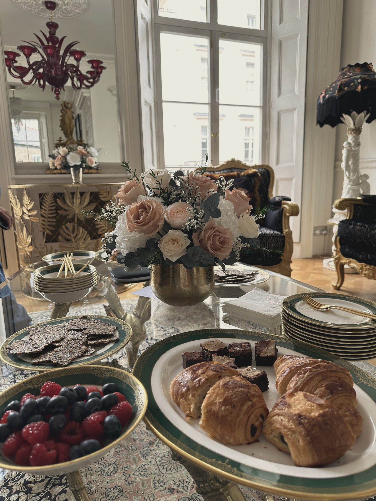 A salon table laid: roses in a brass bowl, gold-rimmed porcelain, pastries and berries, daylight from tall windows