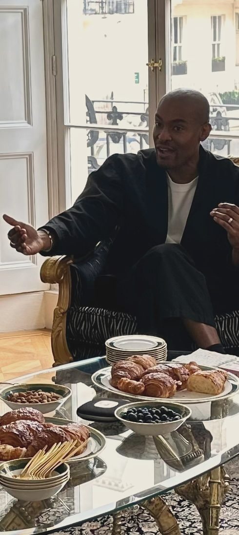 A guest expert mid-conversation at a salon, morning light through tall windows behind him, breakfast pastries laid on the table in front