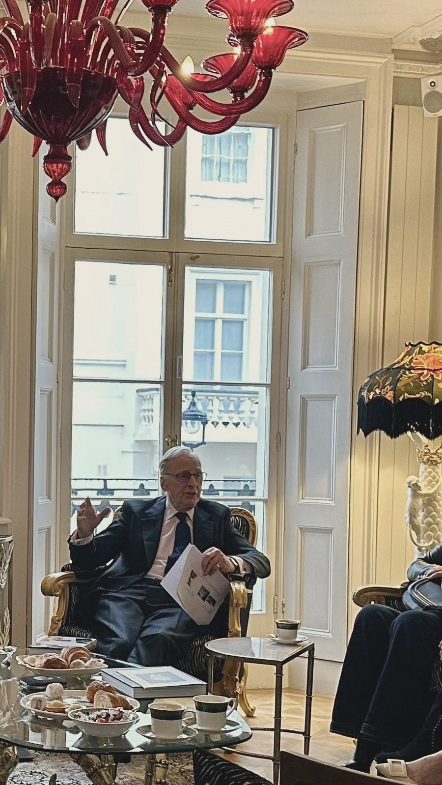 A guest expert mid-sentence at a salon in a London drawing room, tall windows behind him, tea and pastries laid on the table in front