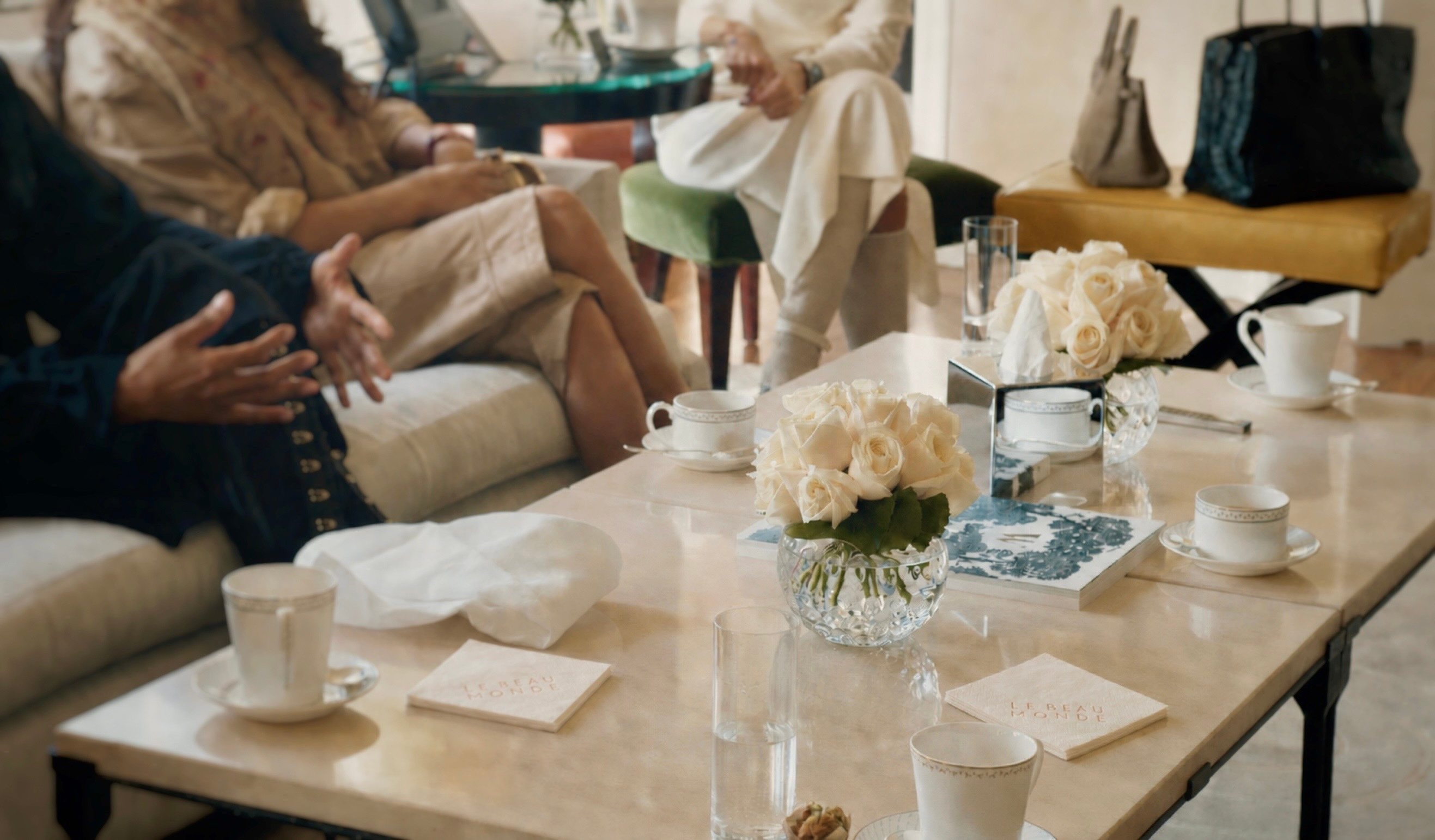 A salon in progress: hands mid-conversation, teacups, white roses and embossed napkins on marble