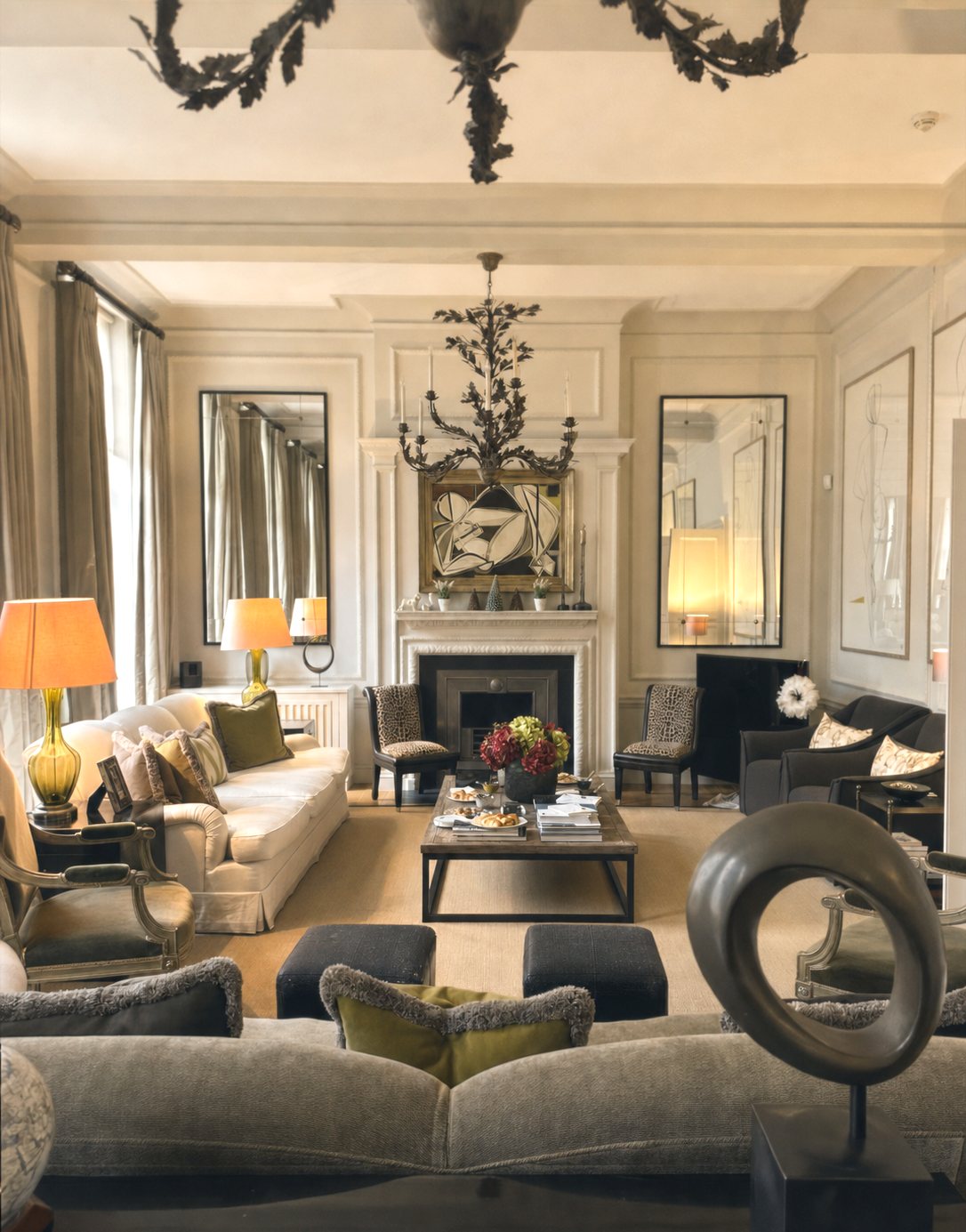 A formal drawing room at Le Beau Monde: a wrought-iron chandelier, a marble fireplace with framed art above it, cream and grey sofas, warm lamplight, no one present